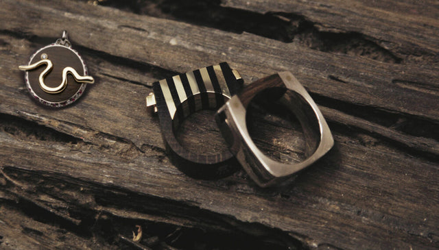 three pieces made with thames oak lying on top of a bigger piece of thames oak. There is a charm with a gold inlaid river thames, and two rings made with gold and silver and thames wood. The bridge of sighs ring and the tenon ring.